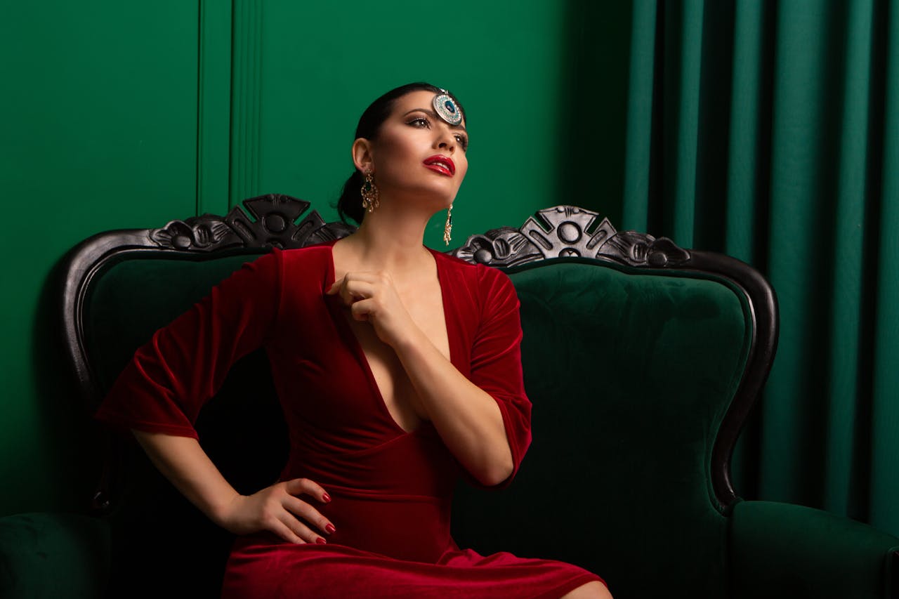 Woman in a red dress and maang tikka posing elegantly on a green settee indoors.